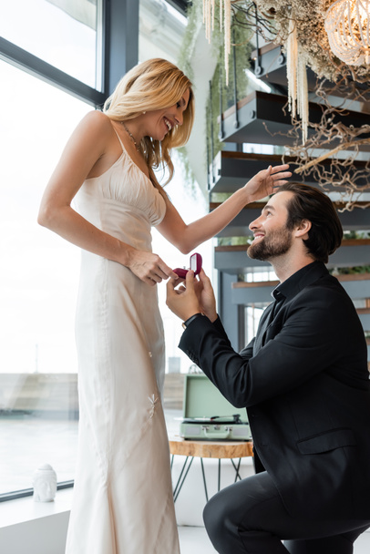 Side view of man holding box with engagement ring near cheerful girlfriend in restaurant  - Valokuva, kuva