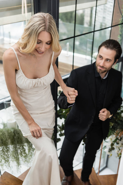 Smiling woman in dress holding hand of blurred boyfriend on stairs in restaurant  - Foto, Bild