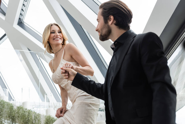 Low angle view of smiling woman in dress holding hand of blurred boyfriend in restaurant  - Foto, Imagen