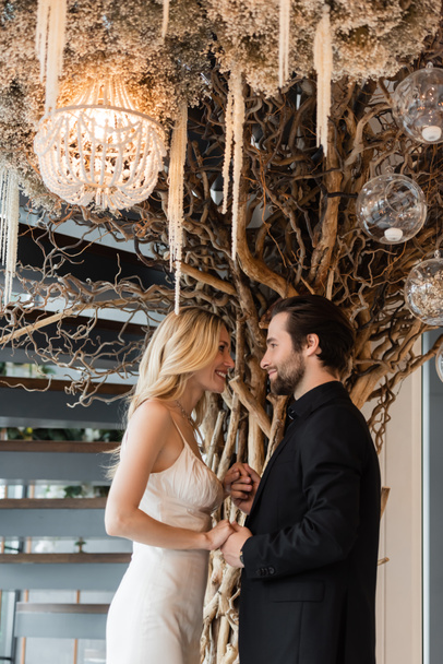 Вид сбоку улыбающейся элегантной пары, держащейся за руки возле декоративной елки в ресторане  - Фото, изображение