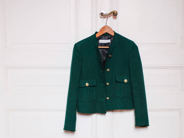 Stylish women's trendy green jacket with gold large buttons hangs on a wooden coat hanger over the doorknob of an antique white door with little copy space, close-up side view. The concept of fashion accessories. - Valokuva, kuva