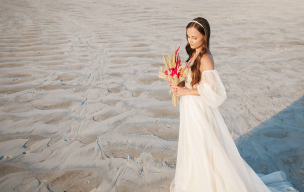 Красивая девушка в свадебном платье позирует перед пустыней, песком, каньоном с букетом в руках - Фото, изображение