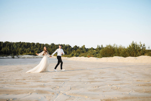 Happy newlyweds are running on the sand against the background of the forest, holding hands. - Fotografie, Obrázek