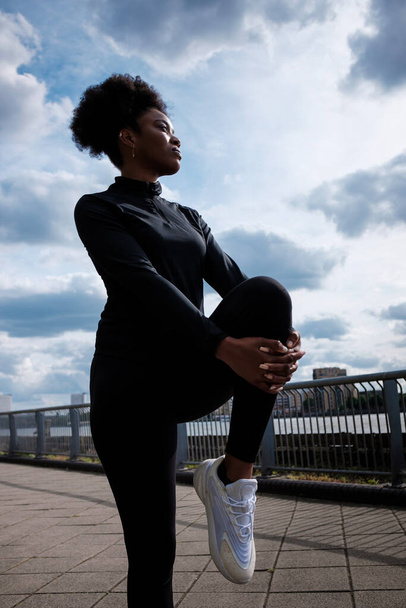 A fit black young woman is stretching close to river Thames in London. He is wearing black sport wear, curly hair. - Foto, imagen