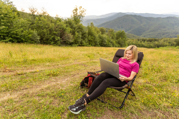 A woman in a sports warm suit works on a laptop outdoors in a mountainous area - Φωτογραφία, εικόνα
