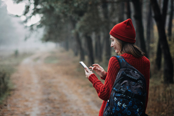 Travel Apps. Young woman traveler with backpack holding smartphone device in hands while walking in pine forest. Happy woman enjoying new adventure and using travel apps or gps navigation in cell - Photo, Image