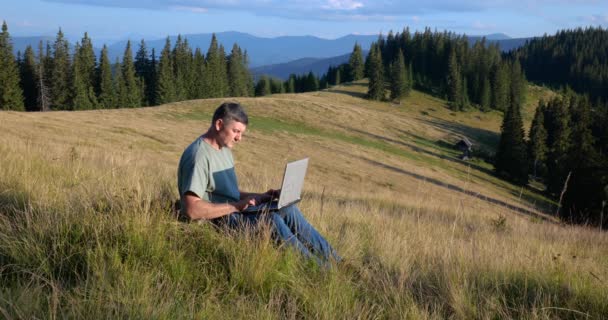 A man sits on a beautiful meadow in the mountains, works on a laptop. Concept of freelancing, digital nomad or remote office. - Filmmaterial, Video