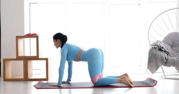 Young fit woman in sportswear doing fitness aerobic exercises with elastic band in living room. Home fitness and wellness concept. - 写真・画像
