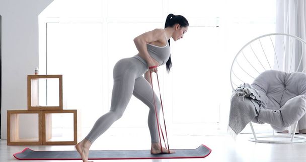 Young fit woman in sportswear doing fitness aerobic exercises with elastic band in living room. Home fitness and wellness concept. - Foto, afbeelding