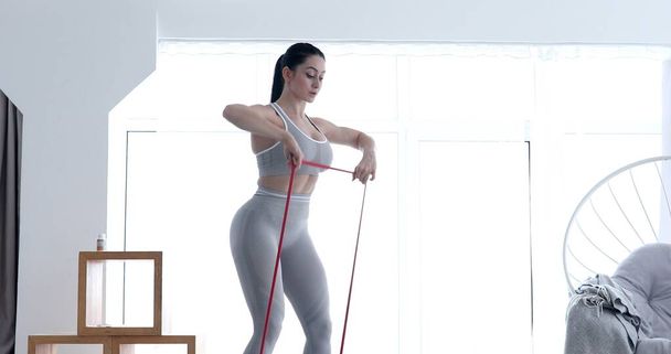 Young fit woman in sportswear doing fitness aerobic exercises with elastic band in living room. Home fitness and wellness concept. - Fotó, kép