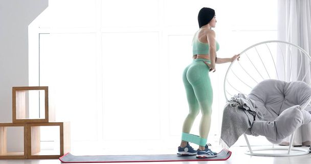 Young fit woman in sportswear doing fitness aerobic exercises with elastic band in living room. Home fitness and wellness concept. - Φωτογραφία, εικόνα