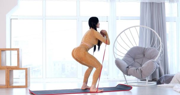 Young fit woman in sportswear doing fitness aerobic exercises with elastic band in living room. Home fitness and wellness concept. - Fotografie, Obrázek