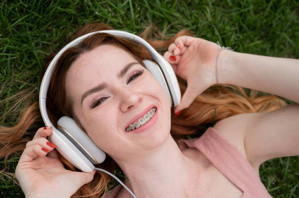 A young red-haired woman lies on the grass and listens to music - Photo, Image