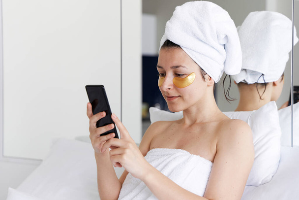 After a shower, the girl is wrapped in a towel and wears cosmetic patches for the skin under the eyes. Looks at the cell phone. Cosmetic procedures at home. Getting ready for a date. - Фото, изображение