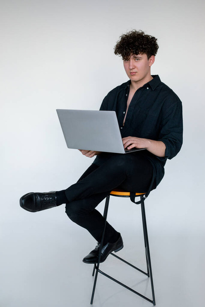 Full length photo of tired young curly guy in total black look sitting cross legged on chair, typing and working on modern laptop, gray studio background. Modern Internet reality, online worker. - Fotoğraf, Görsel