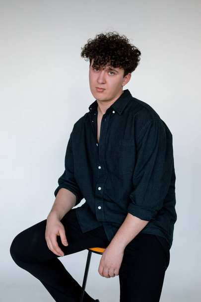 Beautiful young curly guy in total black look sitting on chair, gray studio background. Trendy casual clothes for men, basic shirt. Comfortable fashion wear for every day, street style clothing. - Foto, Imagen