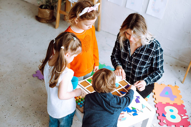 Top view of group of preschoolers and teacher. Children standing around table in bright classroom. Young blond woman showing how to play board games, collecting geometric figures. Preschool education. - Фото, изображение