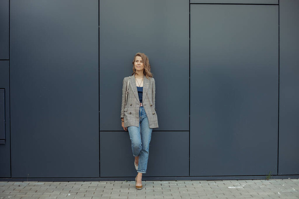 Full length φωτογραφία της σκέψης νεαρή επιχειρηματίας στέκεται στο ένα πόδι ακουμπά στο γκρίζο τοίχο του κτιρίου γραφείων και κοιτάζοντας προς τα πάνω. Αστικός τρόπος ζωής, επιχειρηματίας, καλοντυμένη κυρία, επιτυχημένοι άνθρωποι. - Φωτογραφία, εικόνα