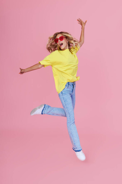 Full length portrait of a joyful young woman in casual clothes jumping and celebrating on a pink background - 写真・画像