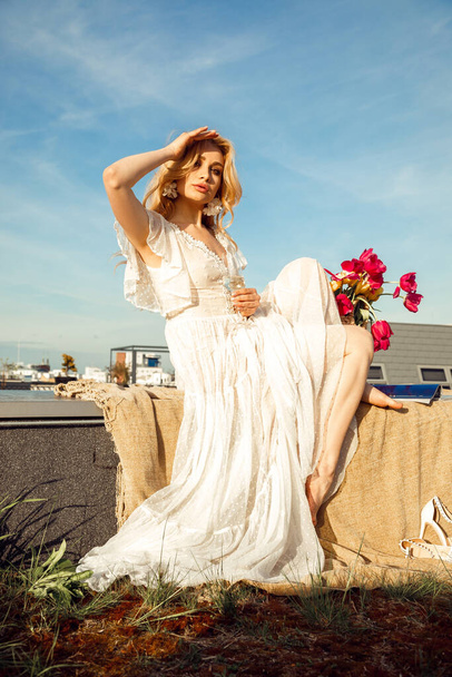 Beautiful young girl in romantic dress with glass of drink covering from sun and sitting on terrace with basket of fresh pink tulips. Paris style, relaxation on roof, cityscape, sky, stylish decor.  - Valokuva, kuva