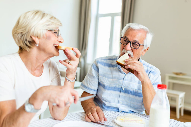 Senior couple eating breakfast at home. Holding piece of bread. Elderly couple enjoy in their time together. Feeling happy. Senior man eating sandwich - Foto, Imagem