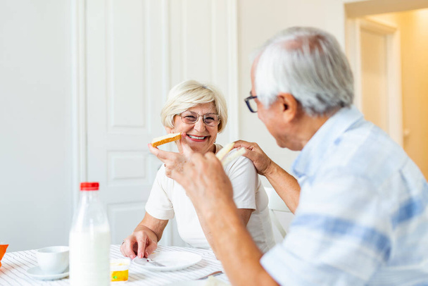 Cheerful senior couple having breakfast at home. Elderly man hugging woman. Mature woman holding piece of bread. An old man and woman sitting at the table, talking. - Photo, image