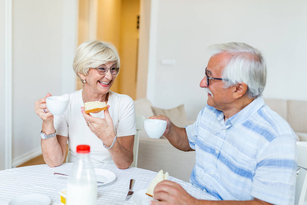 Ηλικιωμένο ζευγάρι τρώει πρωινό και πίνει καφέ. Ηλικιωμένο ζευγάρι τρώει στο σπίτι. Ώριμη γυναίκα που κρατάει κομμάτι ψωμί. Ένας γέρος και μια γυναίκα κάθονται στο τραπέζι, χαλαρώνοντας. - Φωτογραφία, εικόνα