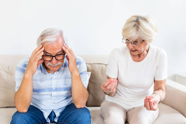 Elderly couple arguing. Senior man having headache, holding his head between hands. Mature woman frustrated with her husband. Marrige problems - 写真・画像