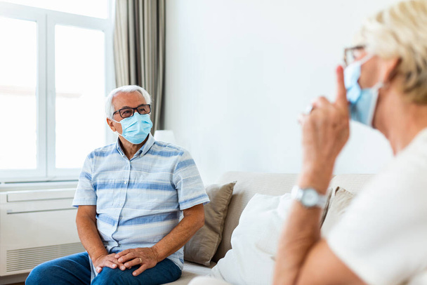 Senior friends having conversation on the sofa at home during Covid 19 pandemic. Elderly wearing medical face mask, preventing covid 19 coronavirus pandemic infection spread. Social distancing - Fotó, kép