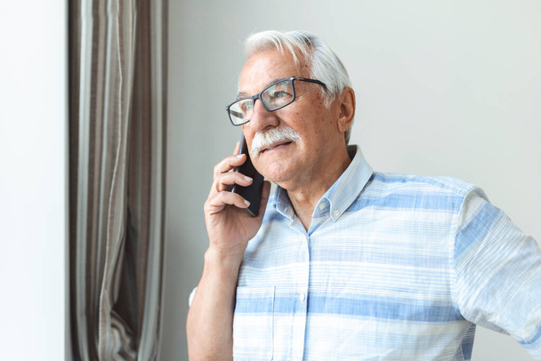 Старший по званию дома разговаривает по телефону. Веселый человек, делающий телефонный звонок и выглядывающий в окно Технология, коммуникация и концепция людей - счастливый пожилой человек звонит на смартфон дома - Фото, изображение