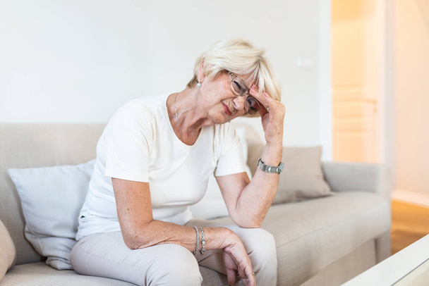 Mature woman sitting on a white sofa in a home touching her head with her hand while having a headache pain and feeling unwell - Foto, afbeelding