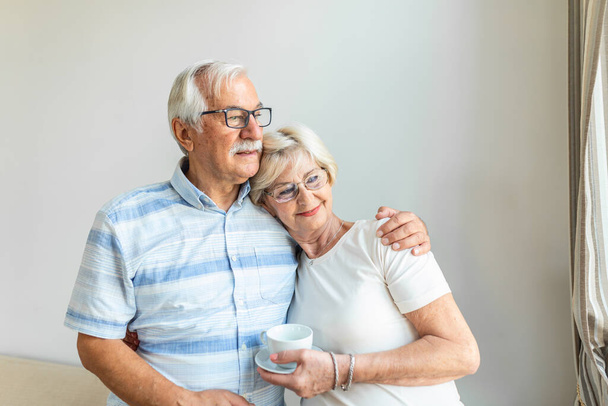Senior couple standing in the living room. Old couple feeling happy,looking in the distance. Cheerful husband hugging his wife. Elderly love concept - 写真・画像