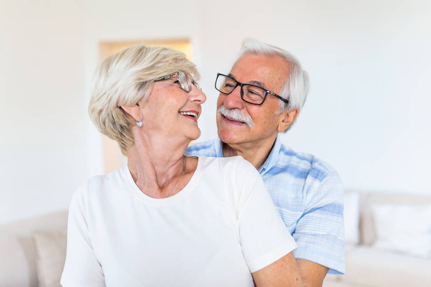 Ηλικιωμένο ζευγάρι στέκεται στο σαλόνι. Πορτρέτο του ευτυχισμένου ηλικιωμένου ζευγαριού αγκαλιάζονται στο σαλόνι στο σπίτι. Το ηλικιωμένο ζευγάρι κοιτάζει ο ένας τον άλλον με αγάπη και χαμόγελο. - Φωτογραφία, εικόνα
