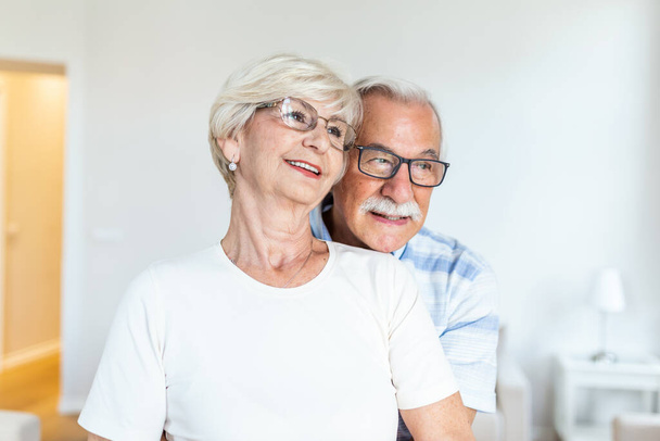 Το ηλικιωμένο ζευγάρι στέκεται στο σαλόνι. Το ηλικιωμένο ζευγάρι αισθάνεται ευτυχισμένο, κοιτάζοντας από απόσταση. Χαρούμενος σύζυγος αγκαλιάζει τη γυναίκα του. Έννοια αγάπης ηλικιωμένων - Φωτογραφία, εικόνα