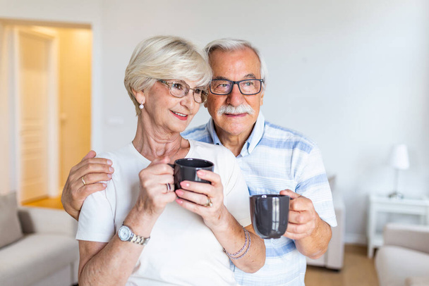 Happy senior couple standing in the living room. Looking each other smilling and holding cup of coffee or tea. Elderly husband hugging his lovely wife - 写真・画像