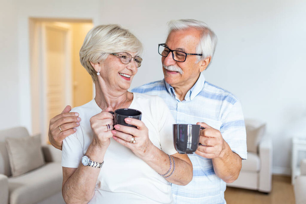 Ευτυχισμένο ζευγάρι ηλικιωμένων στέκεται στο σαλόνι. Κοιτάζοντας ο ένας τον άλλον χαμογελώντας και κρατώντας ένα φλιτζάνι καφέ ή τσάι. Ηλικιωμένος άντρας αγκαλιάζει την όμορφη γυναίκα του - Φωτογραφία, εικόνα