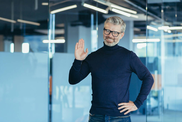 Portrait of successful financier in modern office, man looking at camera and smiling holding hand up greeting gesture, gray haired senior experienced businessman with beard and glasses, investor owner - Photo, image