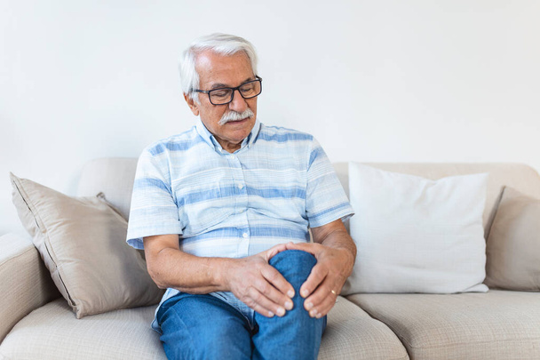 Пожилой мужчина сидит дома на диване и трогает больное колено. Люди, здравоохранение и концепция проблем - несчастный пожилой человек, страдающий от боли в коленях дома - Фото, изображение