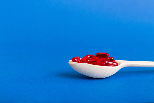Vitamin capsules in a spoon on a colored background. Pills served as a healthy meal. Red soft gel vitamin supplement capsules on spoon. - Foto, Imagem