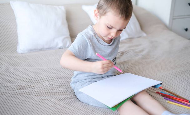 A small caucasian boy in a gray T-shirt and shorts on the bed draws with colored pencils in a sketchbook. Childrens creativity and development. - 写真・画像
