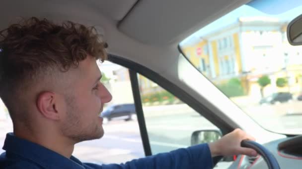 Close up shot of happy young man driving a car in town and smiling. Cheerful positive guy with curly hair drives his auto looking at road. Side view. Caucasian male driver in vehicle goes to work - Footage, Video