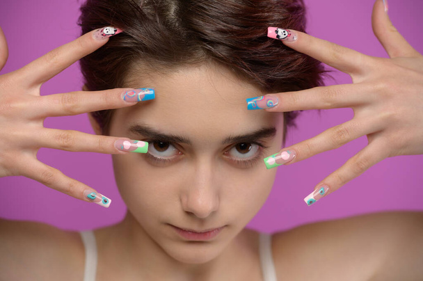 Pretty girl with short haircut and extravagant nail art looking into camera, studio portrait at colored background - Photo, image