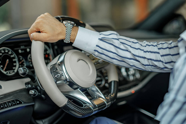 Close up άποψη των αρσενικών χέρι κρατώντας τιμόνι ενός πολυτελούς αυτοκινήτου. Ένας άντρας φοράει ακριβό ρολόι και ριγέ πουκάμισο. - Φωτογραφία, εικόνα