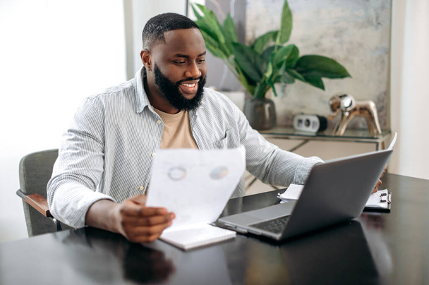 Focused successful positive african american busy man, freelancer or office worker, working documents, sit at a desk in modern office, working on a project uses laptop, studying graphs, smiling - Foto, Imagem