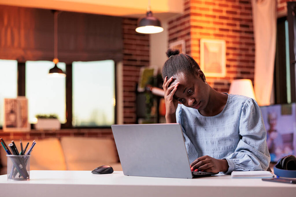 Exhausted female remote worker having headache, young stressed freelancer working on laptop. Unhappy african american employee at workplace in home office with beautiful warm sunset light - Valokuva, kuva