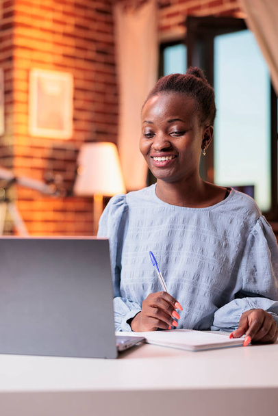 Smiling female student watching educational tutorials on laptop and taking notes in notebook. Young african american woman attending remote university online courses on internet - Photo, Image