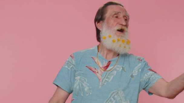 Smiling romantic senior man with flowers in beard sending lots of air kisses looking at camera and smiling, missing you, love confession. Elderly grandfather isolated alone on pink studio background - Video
