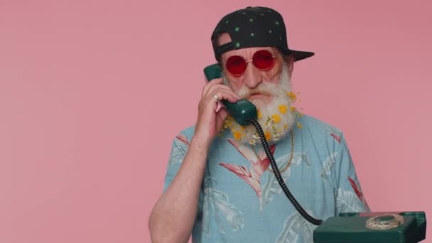 Hey you, call me back. Cheerful bearded old man secretary in t-shirt talking on wired vintage telephone of 60s, asking new client for call. Elderly grandfather isolated on pink studio wall background - 映像、動画