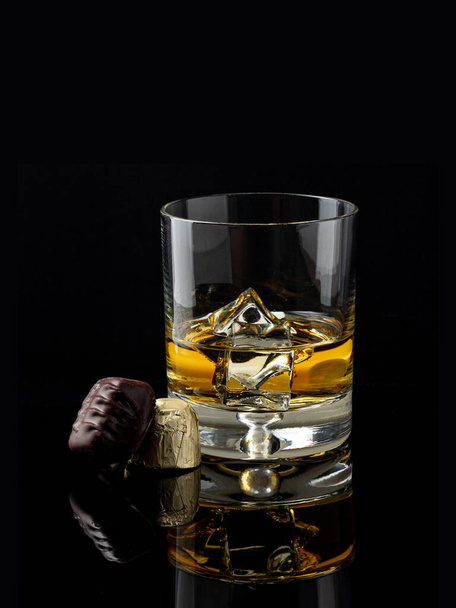 Glass of Kentucky bourbon on ice rocks with two pieces of chocolate candy next to it on black background - Photo, image