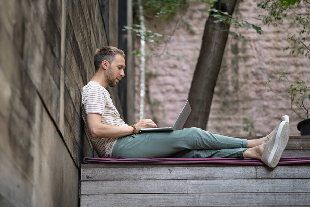 Young European guy, freelancer working on laptop in peace and quiet outside. Focused millennial German man sitting on wooden stairs outdoors in sunshine, studying online at outdoor workspace - Foto, imagen